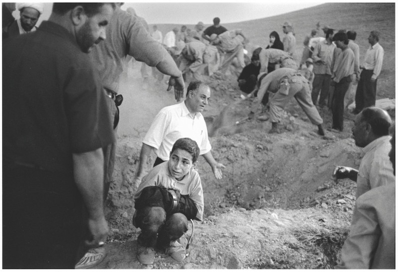 A boy holds his dead father's trousers as he squats beside the spot where his father is to be buried, surrounded by soldiers and villagers digging graves for victims of an earthquake. The earthquake, measuring 6.0 on the Richter scale, struck on June 23. Dozens of villages were destroyed and hundreds of people killed across the province. 23rd June 2002. Qazvin. Iran. Photo Eric Grigorian/Polaris Images 