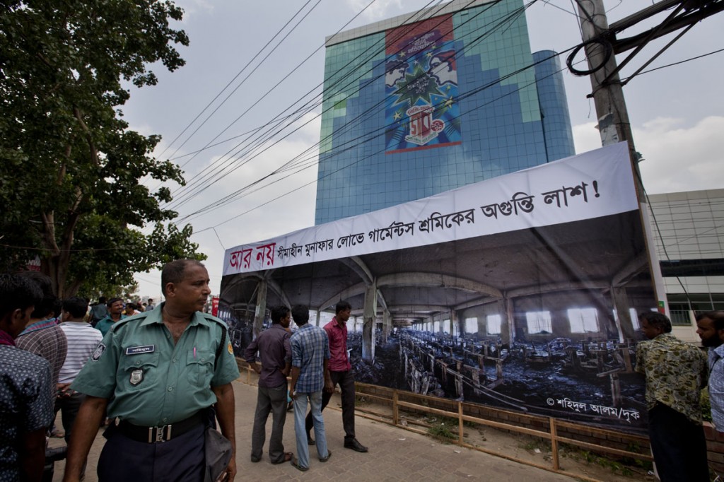 The BGMEA building towers over the 30 foot print of the 3rd floor of Tazreen Fashions. The banner on the building advertises a song contest. A sign, expressing the sadness felt by the owners on the death of workers is much smaller and hidden by the print. The high court has ordered the BGMEA building, built illegally inside a lake, to be taken down, which the BGMEA has appealed against. The sign above the panoramic print. by Drik photographer Shahidul Alam says "NO MORE, innumerable corpses of workers, in the pursuit of unending profits." Photo: Shahidul Alam/Drik/Majority World